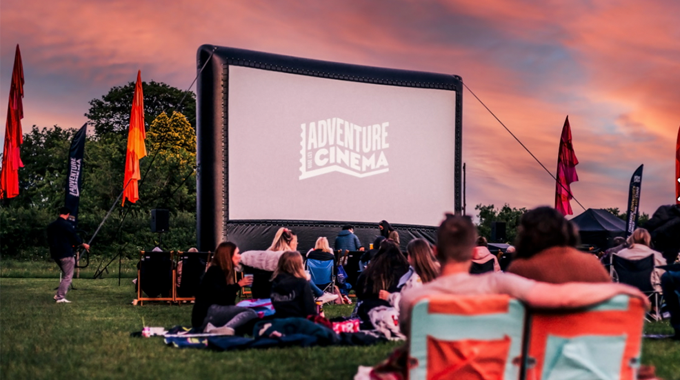 Audience in front of a outdoors cinema screen at sunset with Adventure Cinema logo displayed on screen
